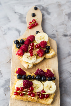 Sendvci with peanut butter, banana and raspberries and currants on a wooden board on a gray table. Vegan healthy breakfastの写真素材