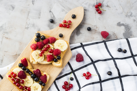 Sendvci with peanut butter, banana and raspberries and currants on a wooden board on a gray table. Vegan healthy breakfastの写真素材