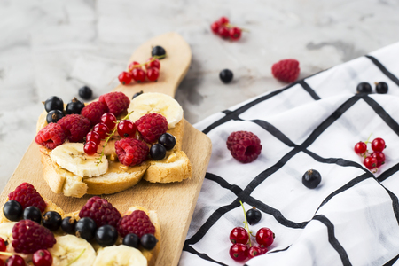 Sendvci with peanut butter, banana and raspberries and currants on a wooden board on a gray table. Vegan healthy breakfastの写真素材