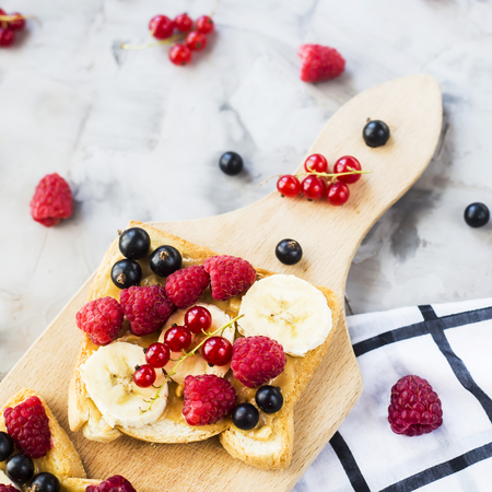 Sendvci with peanut butter, banana and raspberries and currants on a wooden board on a gray table. Vegan healthy breakfastの写真素材