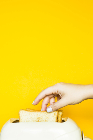 Toasted bread sticks out of the toaster on a yellow background. A woman's hand takes out a piece of bread. Copy spaceの写真素材