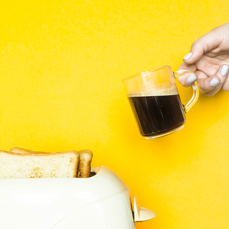 Toasted bread sticks out of the toaster on a yellow background. A female hand holds a cup of coffee. Breakfast conceptの写真素材