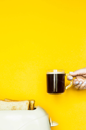 Toasted bread sticks out of the toaster on a yellow background. A female hand holds a cup of coffee. Breakfast conceptの写真素材