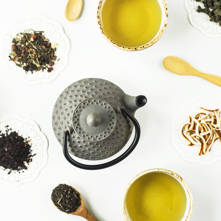 Gray iron kettle among different kinds of dry tea and cups with a ready drink on a white table. Top view, flat layの写真素材
