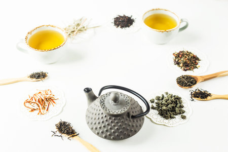 Gray iron kettle among different kinds of dry tea lined up in a circle and cups with a ready drink...