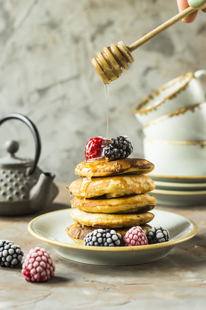 Honey pours on the drain of pancakes on a plate with raspberries and blackberries next to cups and a kettle on a gray table. Summer Breakfast conceptの写真素材
