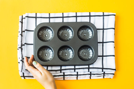 Empty metal mold for baking cupcakes on a black and white checkered napkin with female hand on a yellow background. Top view. Flat layの写真素材