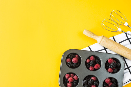 Berries raspberries and blackberries in the form for baking cupcakes on a bright yellow background among kitchen accessories for cooking - rolling pin and whisk of a mixer. Top view, flat layの写真素材