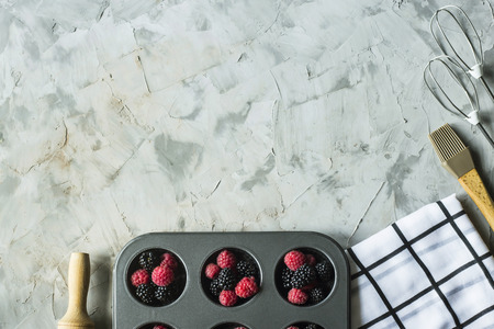 Raspberries and blackberries in metal form for cupcakes next to cooking tools on a gray table. Summer baking concept. Top view, flat layの写真素材