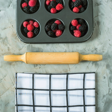 Raspberries and blackberries in metal form for cupcakes next to cooking tools on a gray table....