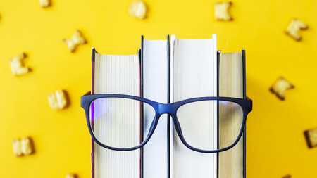 A stack of books with glasses is an image of a person's face. Reading and learning concept. Among the English letters of crackers. On a bright yellow backgroundの写真素材
