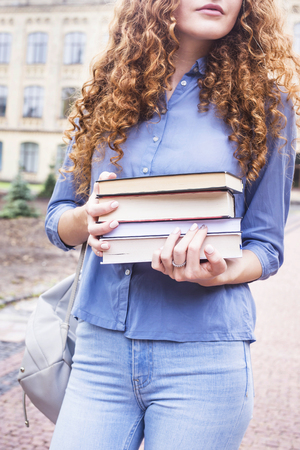 Girl student with long curly red hair with a stack of textbooks from the library. Study at the University of concept. Beginning of the school yearの写真素材