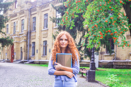 Red-haired girl student with folders and teaching materials in hands next to the university. Back to school concept. student lifeの写真素材