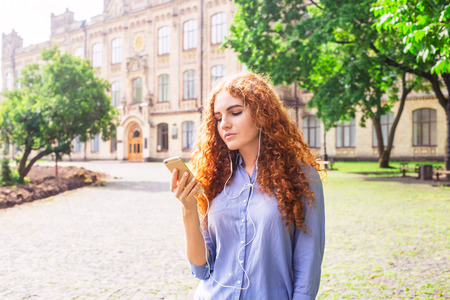 A beautiful young girl with curly red hair looks into her smartphone, to which headphones are connected. Using a smartphone and modern technologyの写真素材