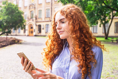 A beautiful young girl with curly red hair looks into her smartphone, to which headphones are...