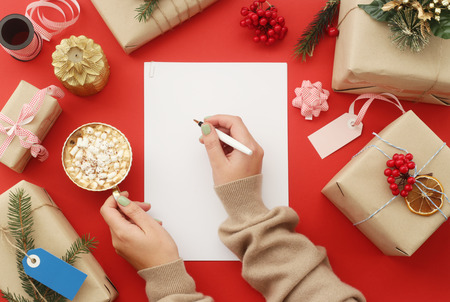Female hand writes on a white sheet of paper among Christmas gifts, decorations next to a cup of cocoa with marshmallows on a red background. Top view, flat layの写真素材