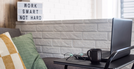 Home workplace of a freelancer at a black stylish table near the sofa and brick wall in a modern apartment. Work smart, no hard - written on a lampの写真素材