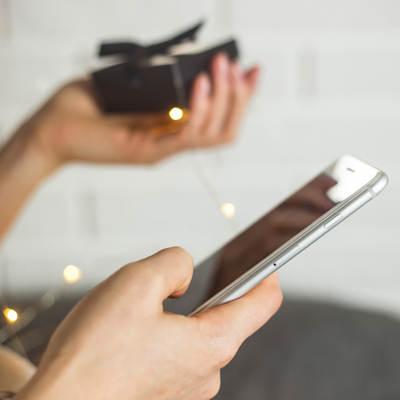 The girl is holding a smartphone with which she orders gifts from online stores from home. Cyber Monday and Black Friday concept. Gift box in hand with lightsの写真素材