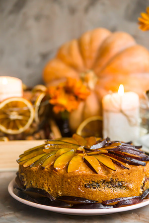Plum cake decorated with sliced plum among the autumn decor - pumpkin, candle, oranges and yellow chrysanthemum. Rustic style. Homely cozy still lifeの写真素材