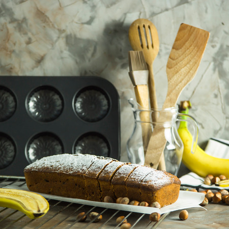 Sliced banana bread is lying on a board among kitchen utensils, bananas and hazelnuts. Homemade bakery conceptの写真素材