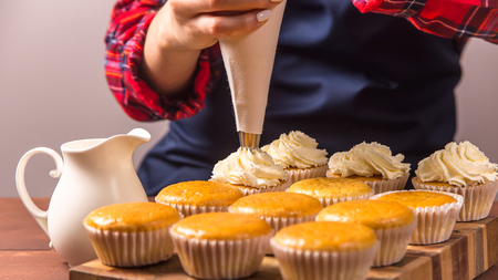 Female confectioner in a blue apron and a plaid red shirt applies cream to cupcakes from a pastry bagの写真素材