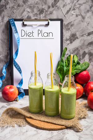 The diet plan is written on a white sheet on the tablet among the bottles of green smoothies, spinach and apples on a gray table with burlap. Weight loss conceptの写真素材