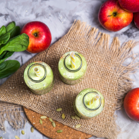 Green smoothies in three glass bottles with tubes on a gray table with a round stand and burlap among apples, spinach and seeds. Top view, flat layの写真素材