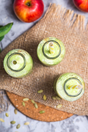 Green smoothies in three glass bottles with tubes on a gray table with a round stand and burlap among apples, spinach and seeds. Top view, flat layの写真素材