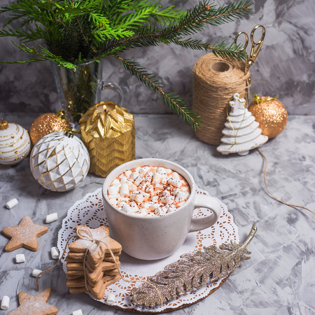 A white cup with hot cocoa with marshmallow stands on a gray table among white and gold Christmas ornaments and fir branchesの写真素材