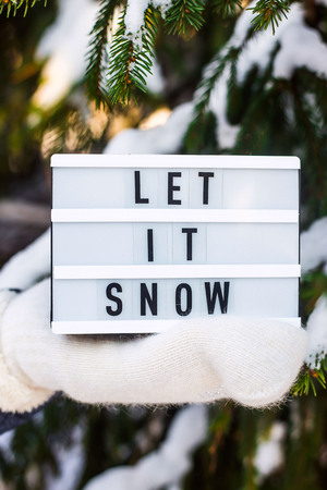 Let it snow it is written on a decorative lamp in the hand of a woman in a white mitten against the background of green spruce branches covered with snowの写真素材