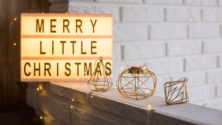 Merry little Christmas is written on a decorative lamp among golden winter decorations and a candlestick on the background of a white brick wall in the apartmentの写真素材