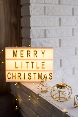 Merry little Christmas is written on a decorative lamp among golden winter decorations and a candlestick on the background of a white brick wall in the apartmentの写真素材