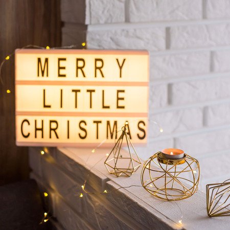 Merry little Christmas is written on a decorative lamp among golden winter decorations and a candlestick on the background of a white brick wall in the apartmentの写真素材