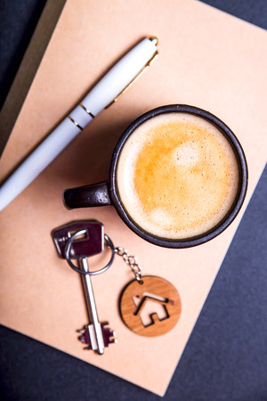 The keys to the new apartment with a wooden key chain house on a black table next to the crafting pad and a cup of coffee. Buying an apartment or house concept. Top view, flat layの写真素材