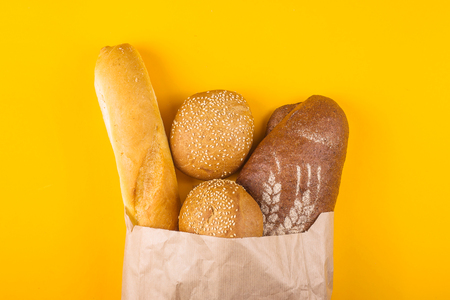 Fresh, varied bread in a paper bag on a yellow background - white baguette, dark loaf and cereal buns with sesame. Top view, flat layの写真素材