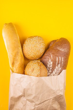 Fresh, varied bread in a paper bag on a yellow background - white baguette, dark loaf and cereal buns with sesame. Top view, flat layの写真素材
