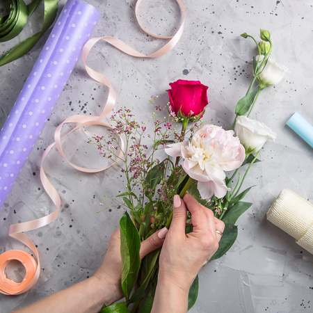 Hands of a woman florist make up a bouquet of white and red roses with gypsophila on a gray table with tools. Top view, flat layの写真素材