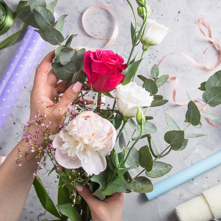 Hands of a woman florist make up a bouquet of white and red roses with gypsophila on a gray table...