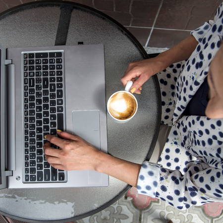 The girl in a light summer jacket sits at a street table cafe with a cup of cappuccino and works at a laptopの写真素材
