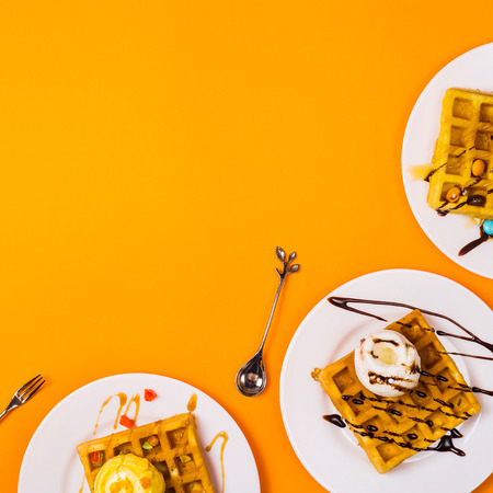 Fresh golden square Belgian waffles on plates with ice cream and toppings on a bright yellow background. Top view, flat lay. Copy spaceの写真素材