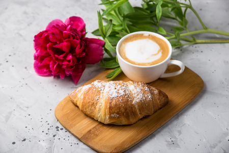 Fresh croissant on a wooden board next to a white cup of cappuccino and red peony on a gray textured tableの写真素材