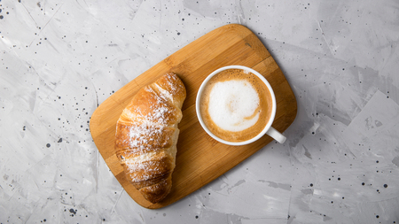 Croissant on a wooden board next to a white cup of cappuccino on a gray textured table. Top view, flat layの写真素材