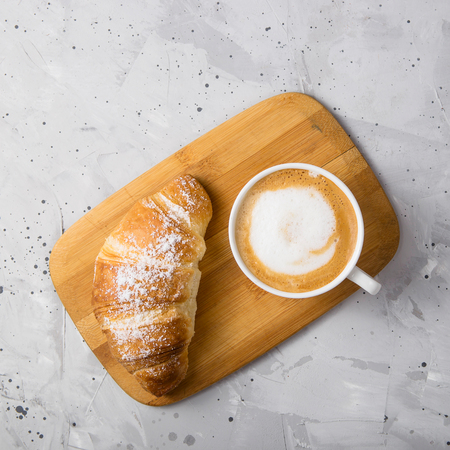 Croissant on a wooden board next to a white cup of cappuccino on a gray textured table. Top view, flat layの写真素材