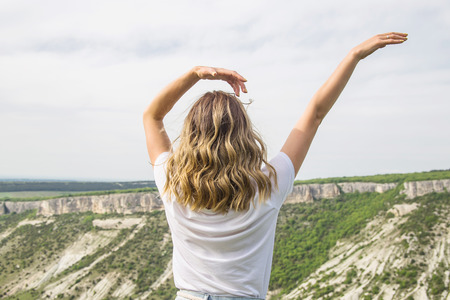 A girl with blond wavy hair in a white T-shirt stands with her back and looks at the mountains with her arms raised. Freedom and travel conceptの写真素材