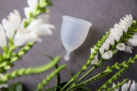 White menstrual cup for women next to white flowers on a gray background. Ecological alternative to gaskets and tampons. Zero waste conceptの写真素材