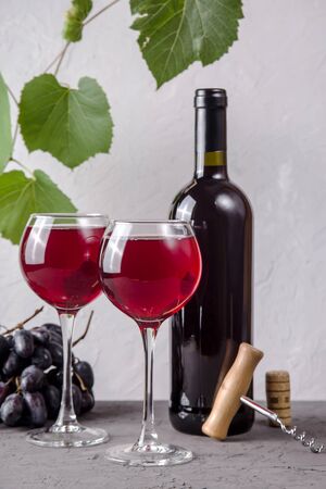 Two glasses of red wine next to a roll, a bunch of grapes and a corkscrew on a background of green branches of grapes on a gray backgroundの写真素材