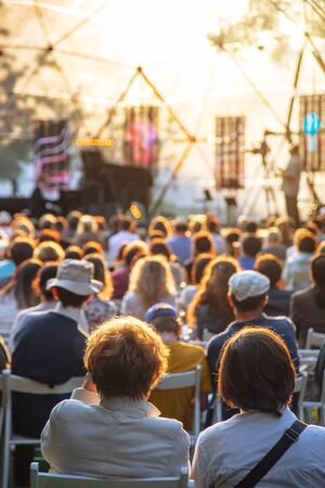 Street concert in the park in summer backs of spectators sitting on chairs at the stageの写真素材