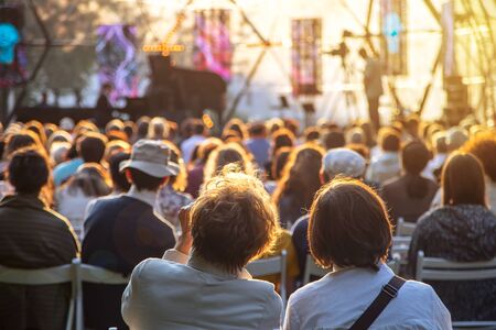 Street concert in the park in summer backs of spectators sitting on chairs at the stageの写真素材