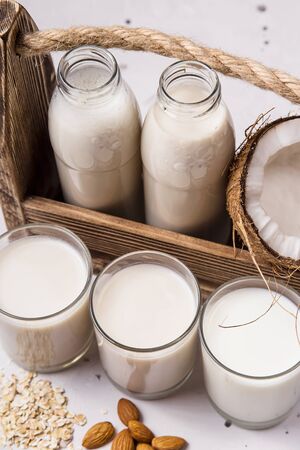 Three alternative vegan milk coconut, oat and almond in glasses next to the ingredients and a wooden box with bottles of milk on a gray tableの写真素材