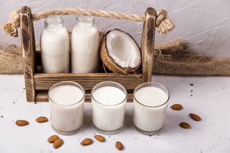 Three alternative vegan milk coconut, oat and almond in glasses next to the ingredients and a wooden box with bottles of milk on a gray tableの写真素材